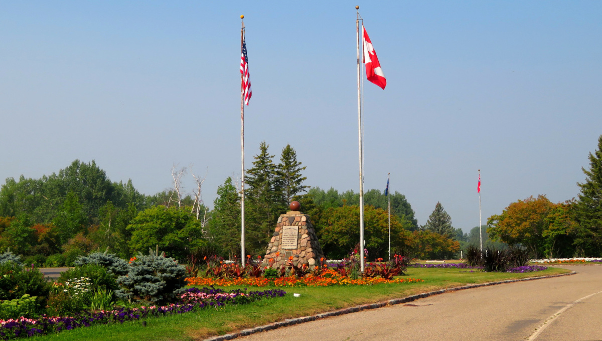 A stone cairn with a carved plaque stands between two flagpoles, one flying the Canadian flag, and the other the flag of the United States.