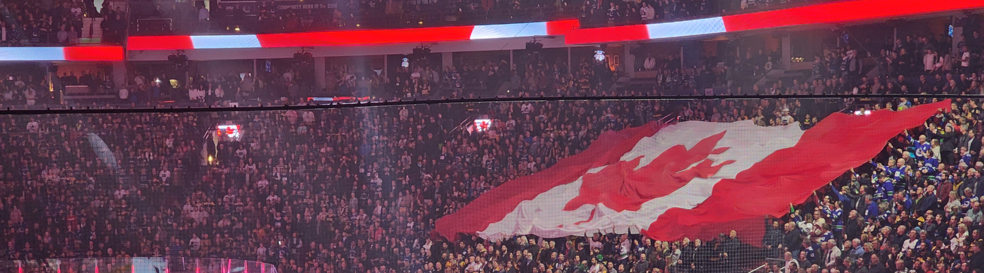An enormous Canadian flag held up by a full stadium audience watching hockey players and referees lined up on the ice.