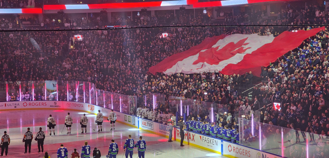 An enormous Canadian flag held up by a full stadium audience watching hockey players and referees lined up on the ice.