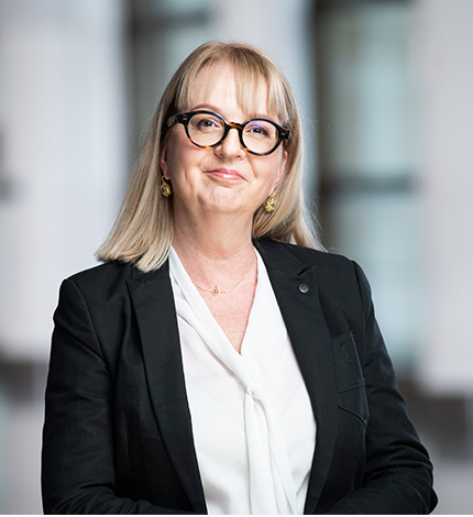 Light-skinned smiling adult with long straight blonde hair, round dark glasses, gold earrings, a dark blazer, and a white blouse.