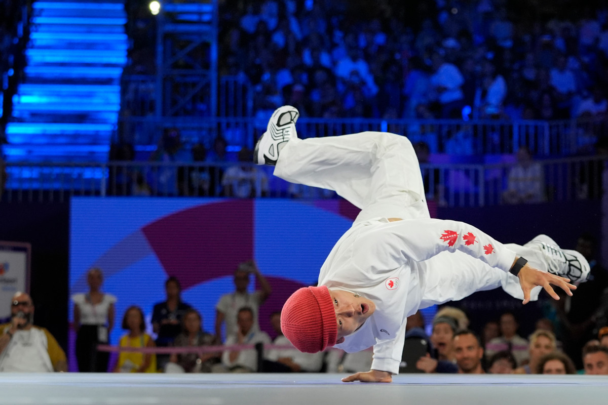 A break dancer with medium skin tone, balancing on one hand with their legs in the air, wearing a red beanie and loose white clothes with three red maple leaves on the forearm.