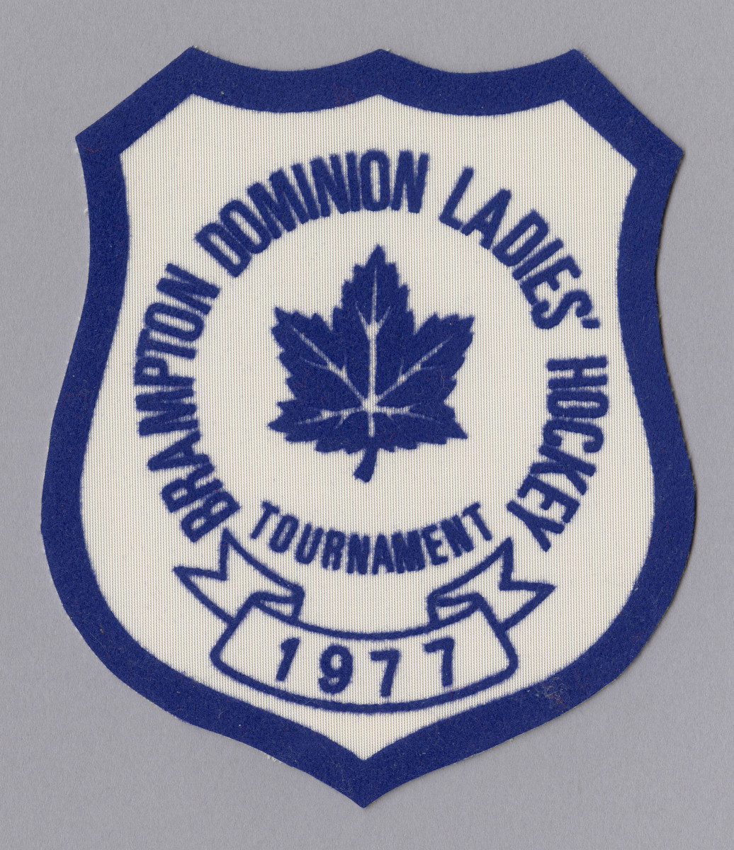 A blue-and-white badge showing a maple leaf surrounded by text reading “Brampton Dominion Ladies’ Hockey Tournament” with “1977” in a banner below.