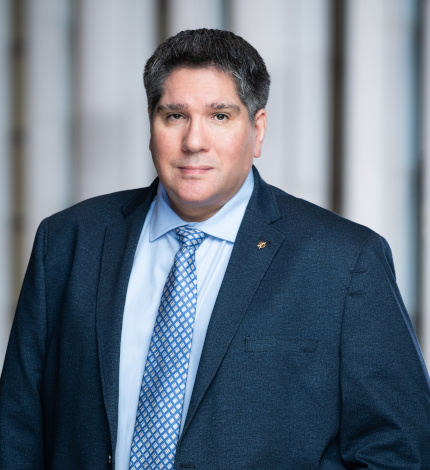Light-skinned adult with short dark hair, a dark blue suit jacket, a light blue dress shirt, and a blue tie.