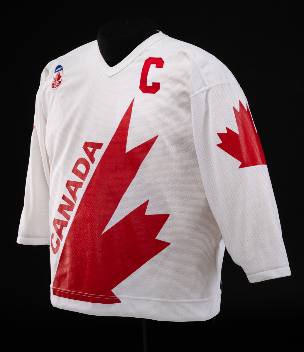 A white hockey jersey with a large, diagonal red maple leaf design and the word “CANADA” on the front, a large “C” on the front left shoulder, and a smaller maple leaf on the arm.
