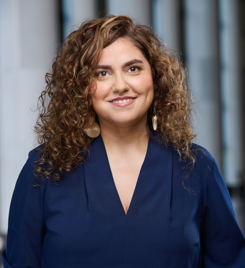 Smiling medium-light-skinned adult with long, curly auburn hair, gold earrings, and a dark blue blouse.