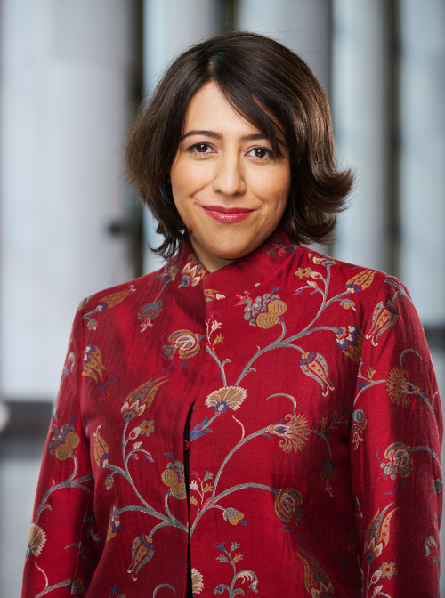 Medum-skinned smiling adult with shoulder-length brown hair and a red silken jacket with floral patterns.
