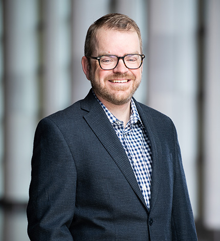 Light-skinned smiling adult with short brown hair, a moustache and beard, glasses, a dark blue jacket, and a checked blue shirt.