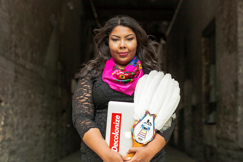 An Indigenous person with long hair, wearing a black lace dress and a pink floral scarf, holding a beaded white ceremonial feather fan and a laptop with a “Decolonize” sticker.