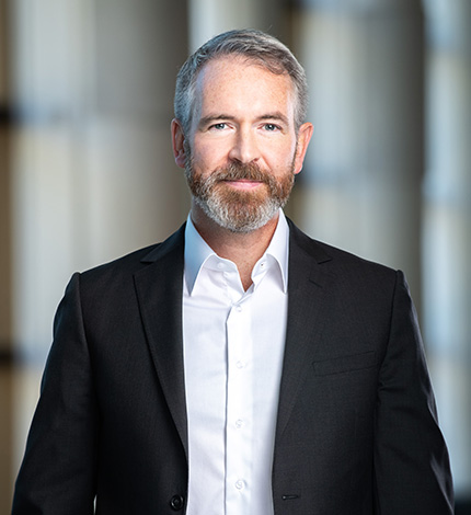 Light-skinned adult with short greying hair, a moustache, and a beard, wearing a black blazer and a white shirt.